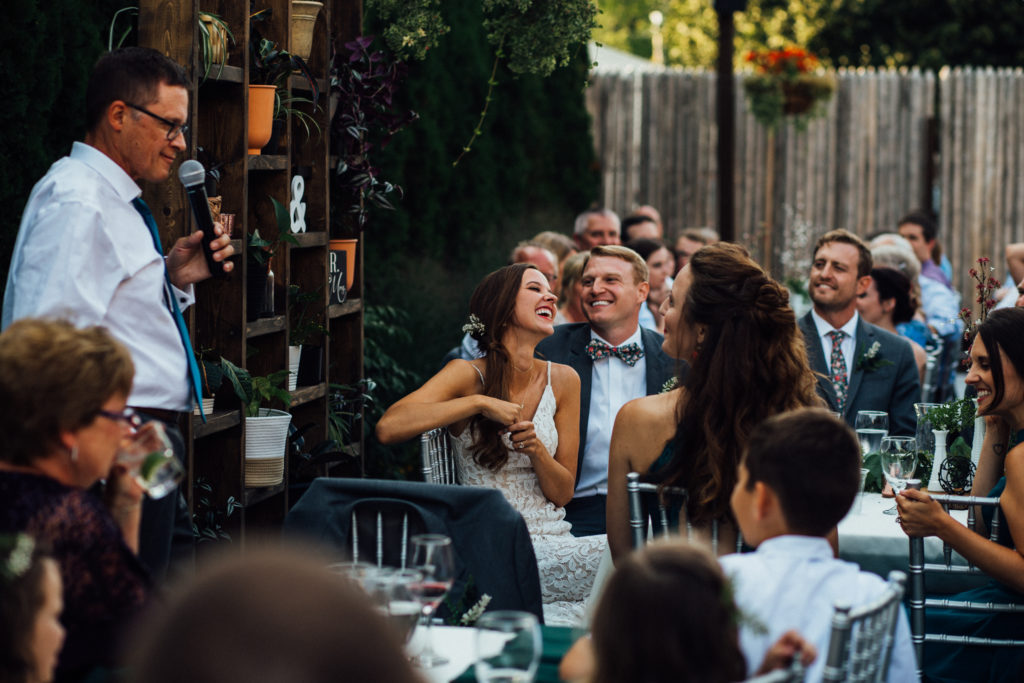 Father of bride speaks during dinner on patio at Birch's wauwatosa outdoor wedding venue while bride and groom laugh