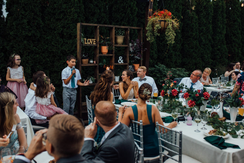 Child giving speech during dinner on patio at Birch while bride and groom laugh