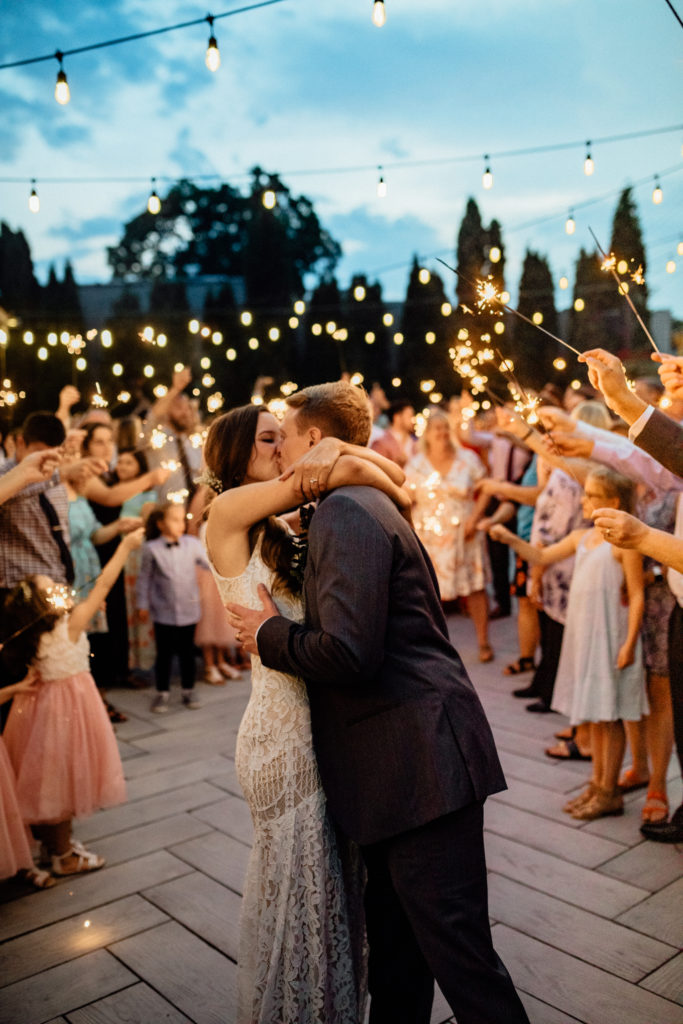 bride and groom embrace in hug and kiss with golden sparklers illuminating them on patio