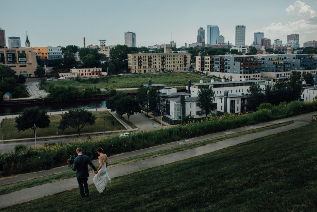 Bride and groom walk down hill at Kadish Park with Milwaukee skyline