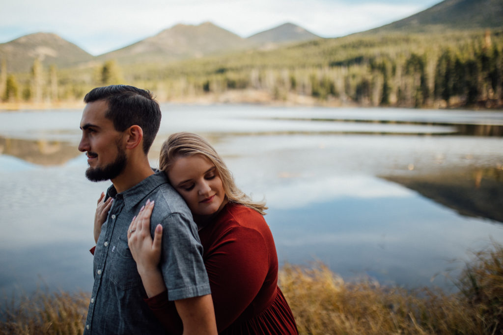 Girl embracing fiance from behind under arms with Sprague Lake in background
