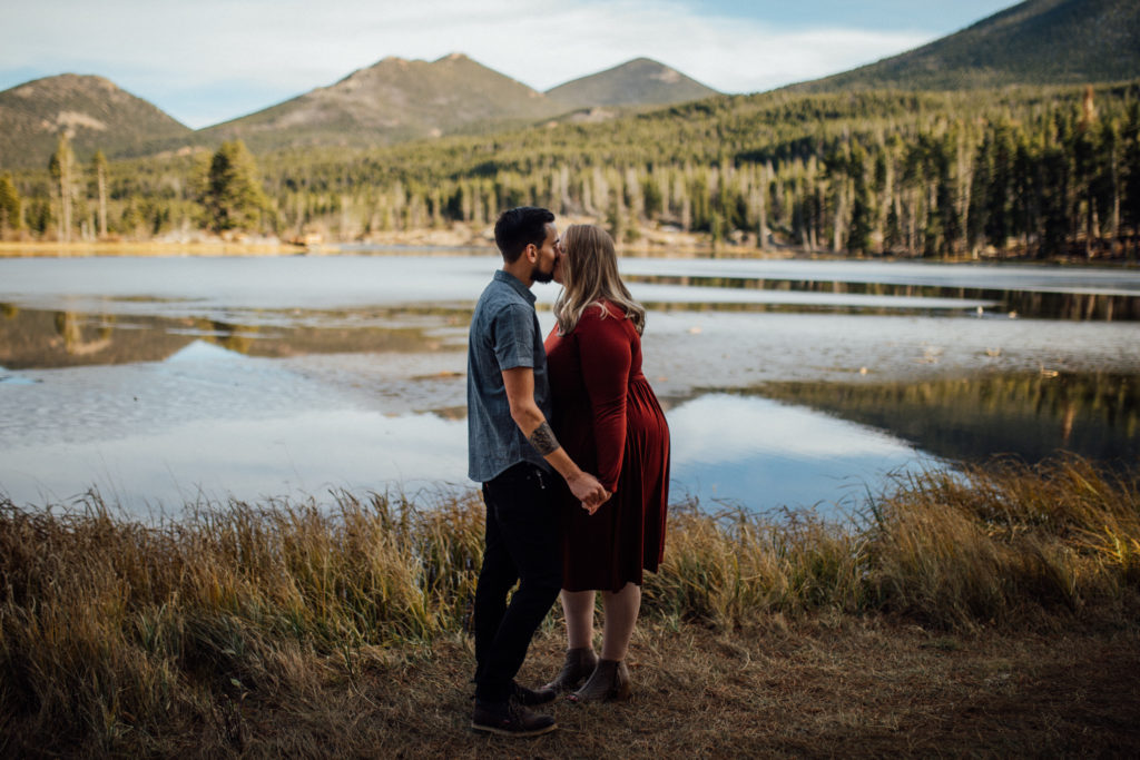 Man and woman kiss in front of Sprague Lake for Rocky Mountain engagement session