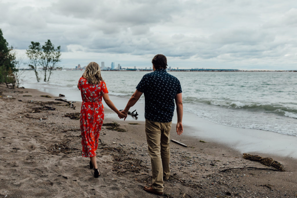 Engaged man and woman walking away on beach towards Milwaukee skyline