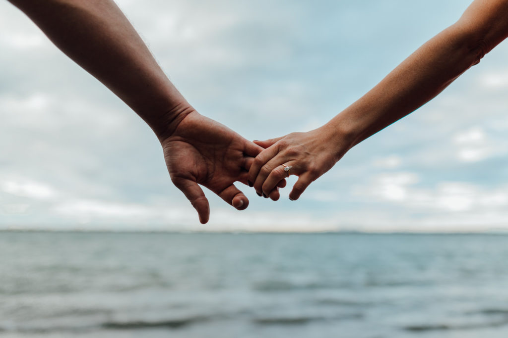 man and woman gently hold hands over horizon of lake with gold teardrop engagement ring