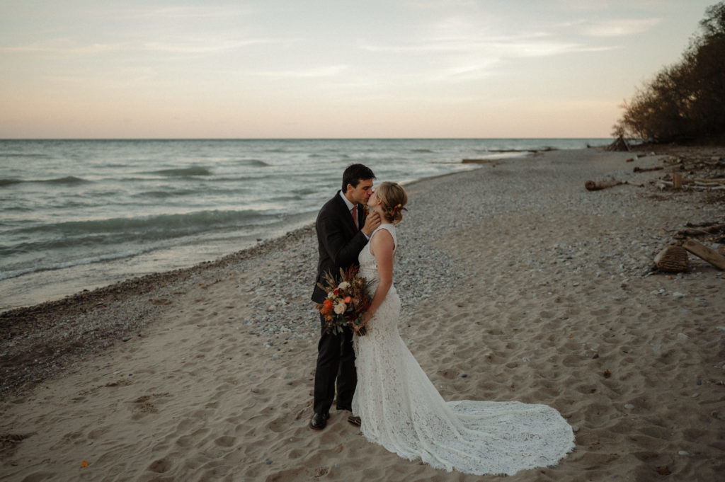 bridge and groom kiss at sunset holding flowers in winter on the best beaches in milwaukee for your engagement session at Doctor's Park