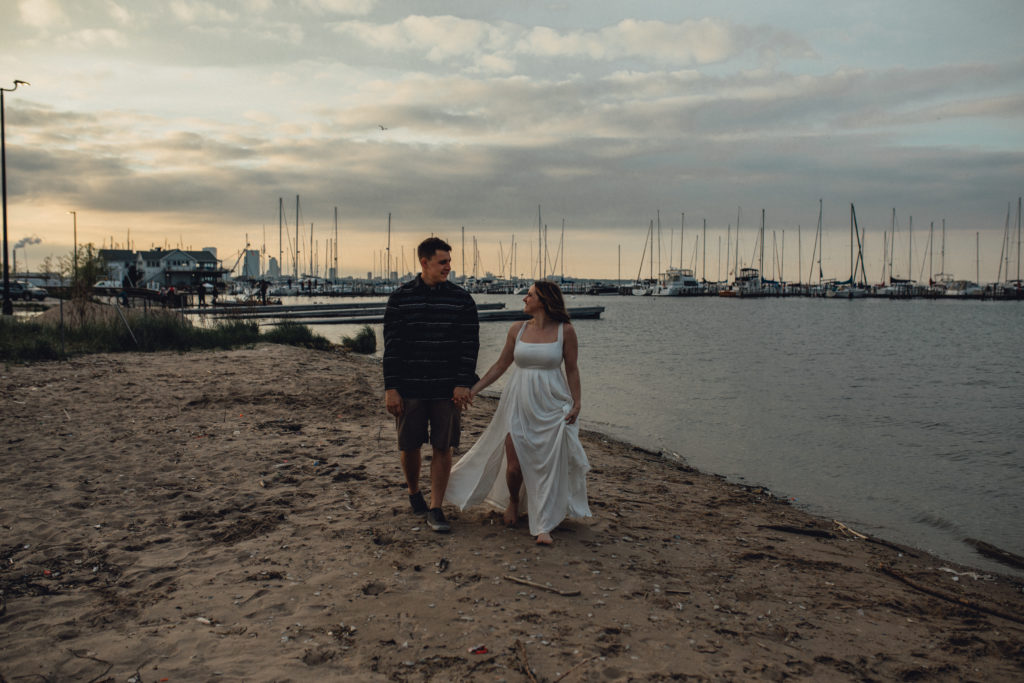 couple walking down south shore beach in milwaukee with boats and marina in the distance at sunset